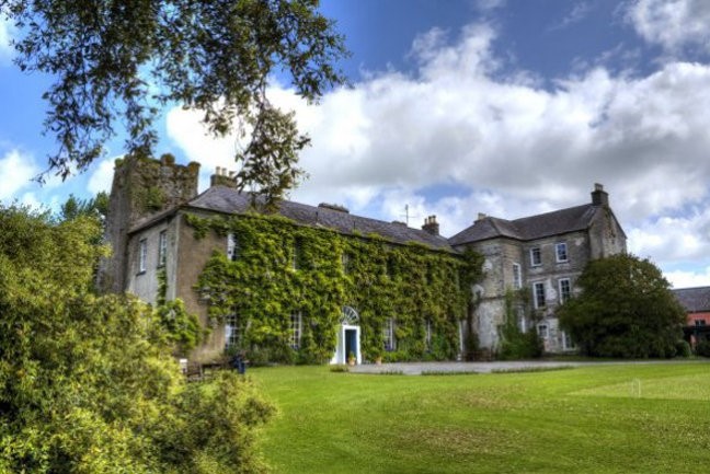Ballymaloe House, een historisch stenen landhuis versierd met groene klimop, ligt tussen weelderige bomen en een keurig onderhouden gazon. Meerdere gevels en schoorstenen rijzen op onder de bewolkte blauwe lucht. Een centrale, wit omrande toegangsdeur is uitnodigend omlijst door gebladerte, wat de charme van een exquis hotel oproept. | Echt Ierland Vakanties